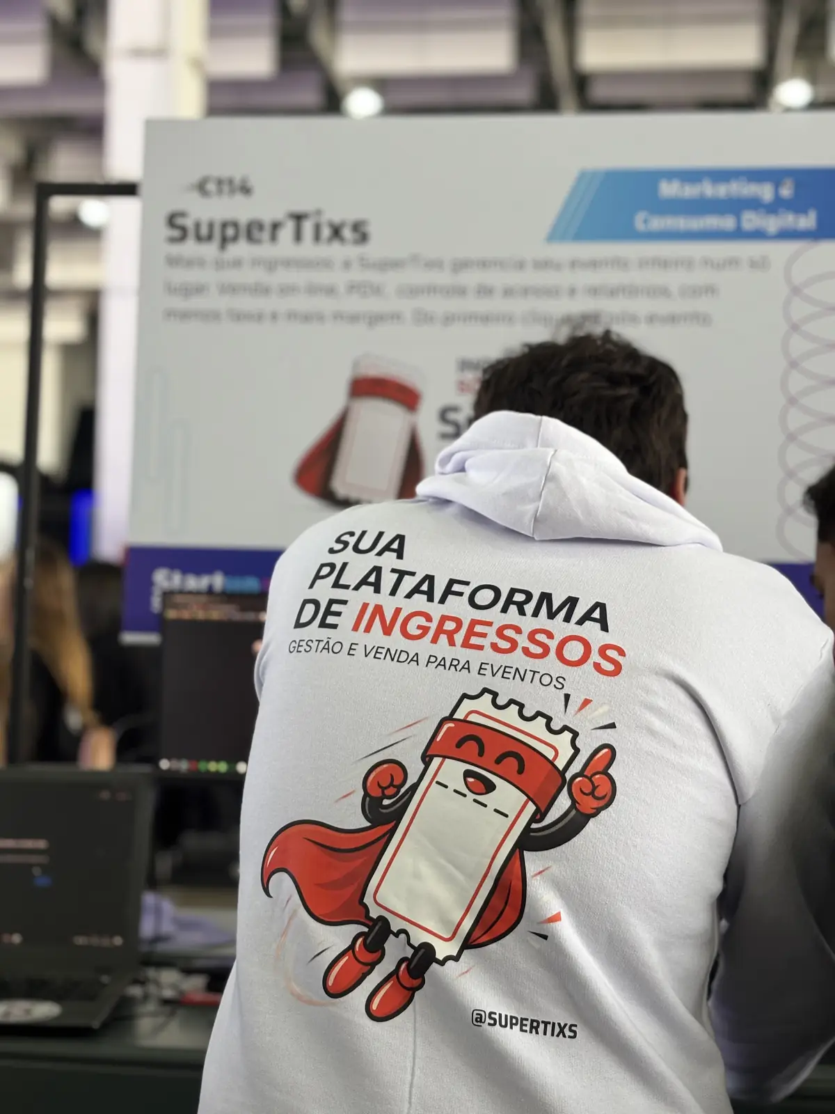 Integrante da equipe de costas, com moletom da SuperTixs e a frase sua plataforma de ingressos